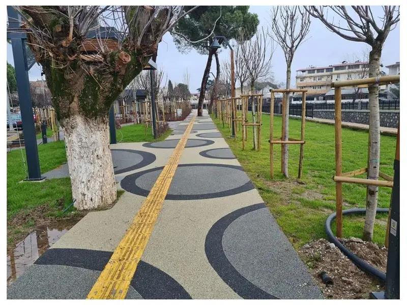 Ağaçlı park içinde agrega taş kaplı yürüyüş yolu ve sarı yönlendirme çizgisi